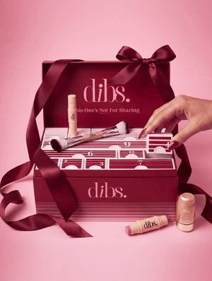 A DIBS cosmetics advent calendar gift set on a pink background, featuring a dark red box with a matching ribbon, numbered compartments, makeup items, and a hand opening one of the compartments.