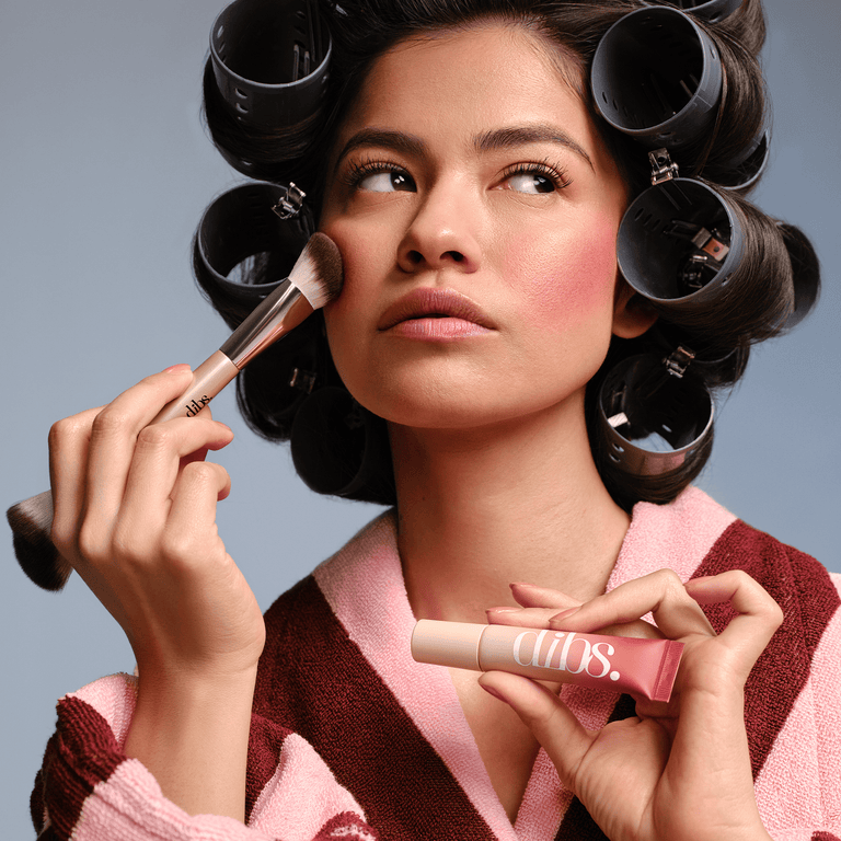 Woman applying liquid blush with a brush, wearing curlers against a gray background