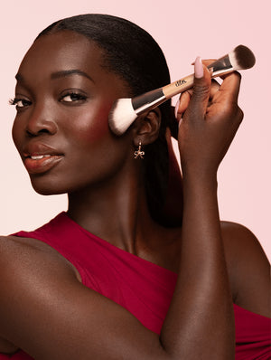 Woman applying makeup to cheek with a double ended makeup brush on a pink background