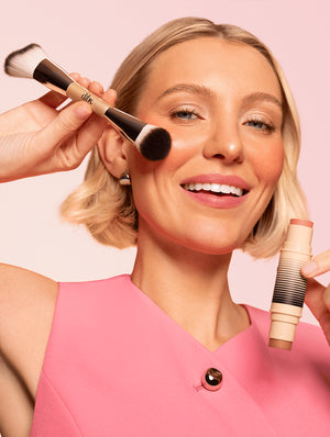Woman applying makeup with a double ended makeup brush and a blush and bronzer makeup stick on a pink background