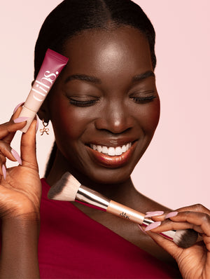 Woman holding a double ended makeup brush and tube of liquid blush with a pink background