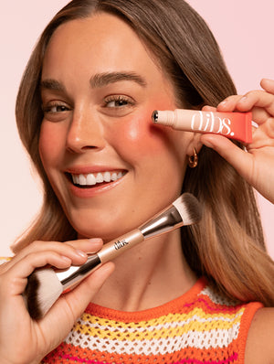 Woman applying liquid blush with a duo ended brush on a pink background