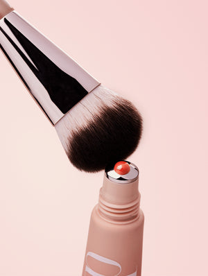 Makeup brush with liquid blush coming out of applicator on a light pink background