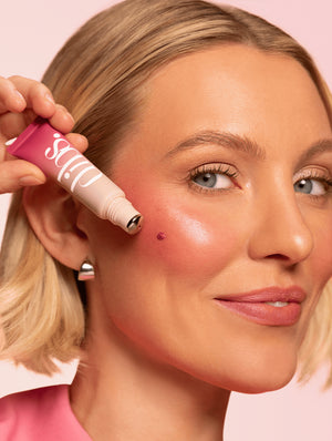 Woman applying a pink liquid blush to her cheek with a pink background