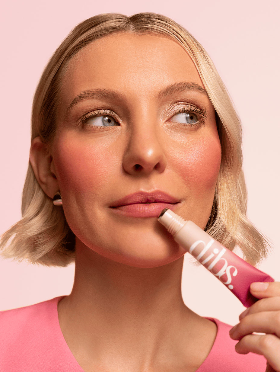 Woman applying a pink liquid blush to her lips with 'dibs' branding on a pink background