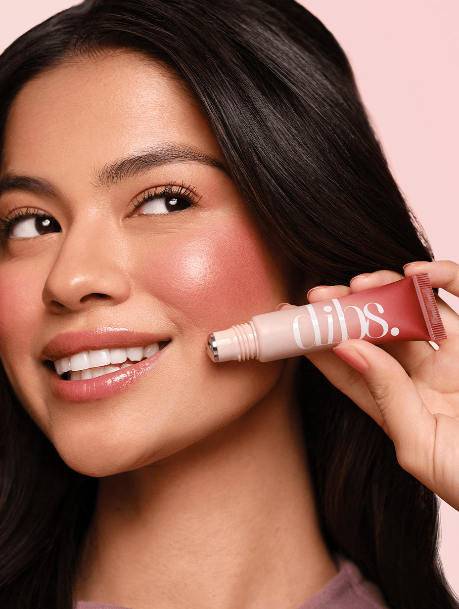 Woman applying liquid blush to her cheek with a 'dibs' packaging on a pink background