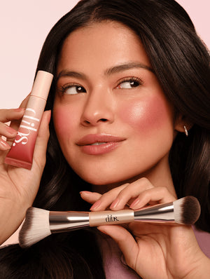 Woman applying bronzed terracotta liquid blush with a brush on a pink background