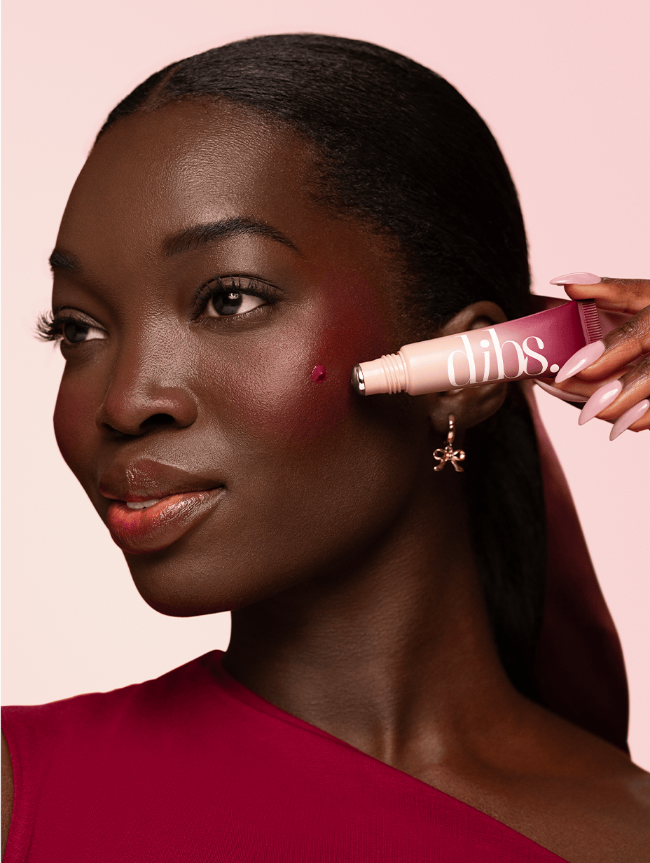 Woman applying liquid blush dot to her face with a pink background