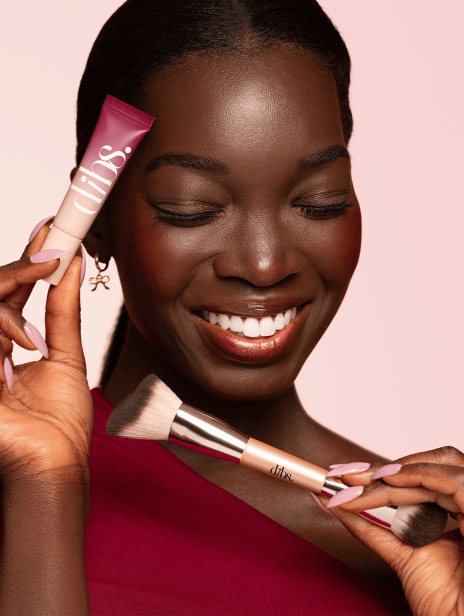 Woman holding a makeup brush and a pink tube of liquid blush with 'dibs' branding on a pink background