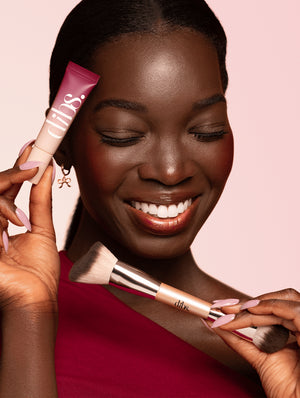 Woman holding a makeup brush and a pink tube of liquid blush with 'dibs' branding on a pink background