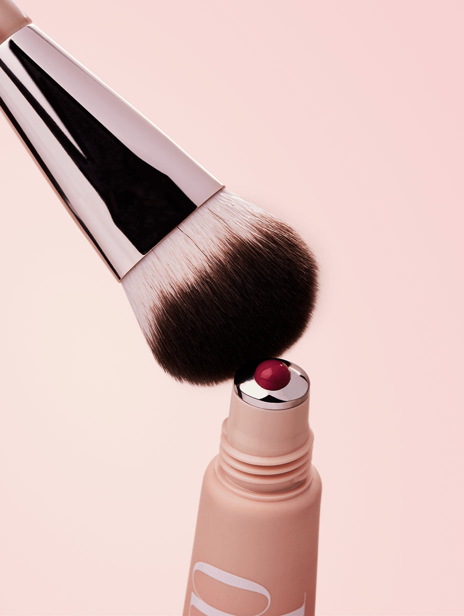 Makeup brush with liquid blush coming out of applicator on a light pink background