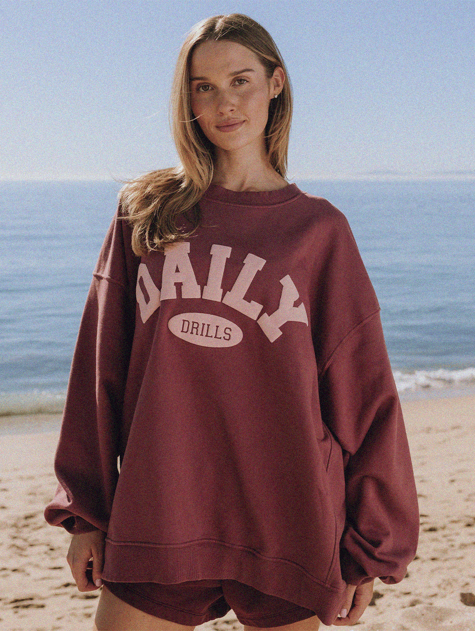 Person wearing a burgundy sweatshirt with 'DAILY DRILLS' text on the front of sweatshirt, standing on a beach